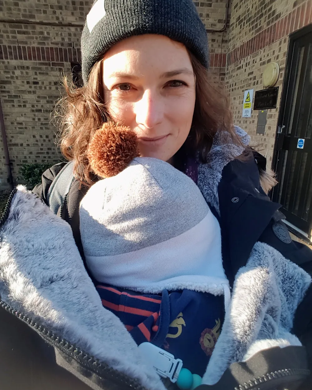 Elodie en bonnet d'hiver, son nouveau-né blotti contre elle en écharpe de portage